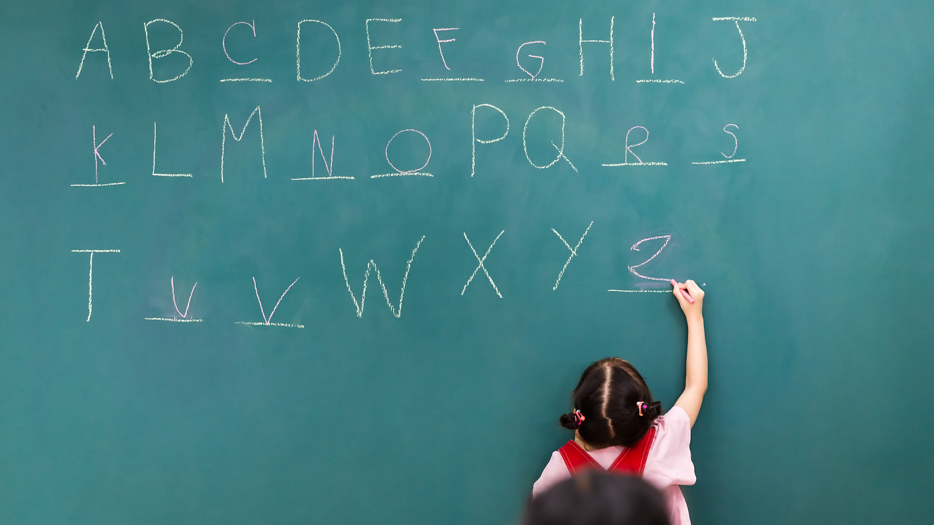 The letters of the alphabet are written out on a chalkboard, but there are blanks in place of a few of the letters. A school girl is writing in the missing letters.